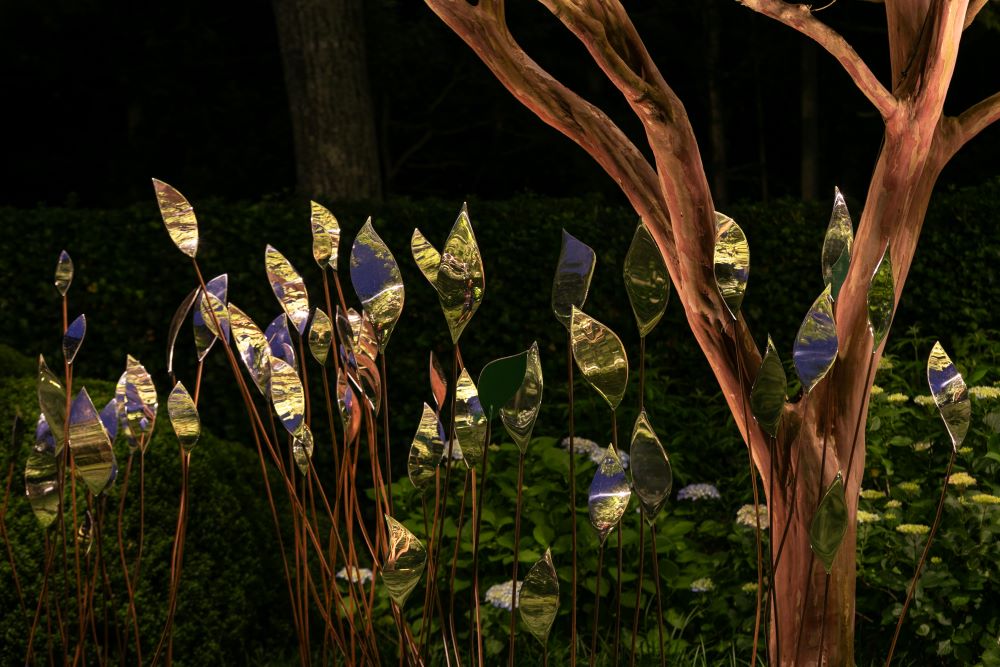 Metallic leaf sculptures, the quiver stainless steel contemporary abstract sculpture, at East Hampton, gleam among real plants and flowers. A twisting tree stands to the right, whilst lush green foliage and dark shadows fill the New York background.