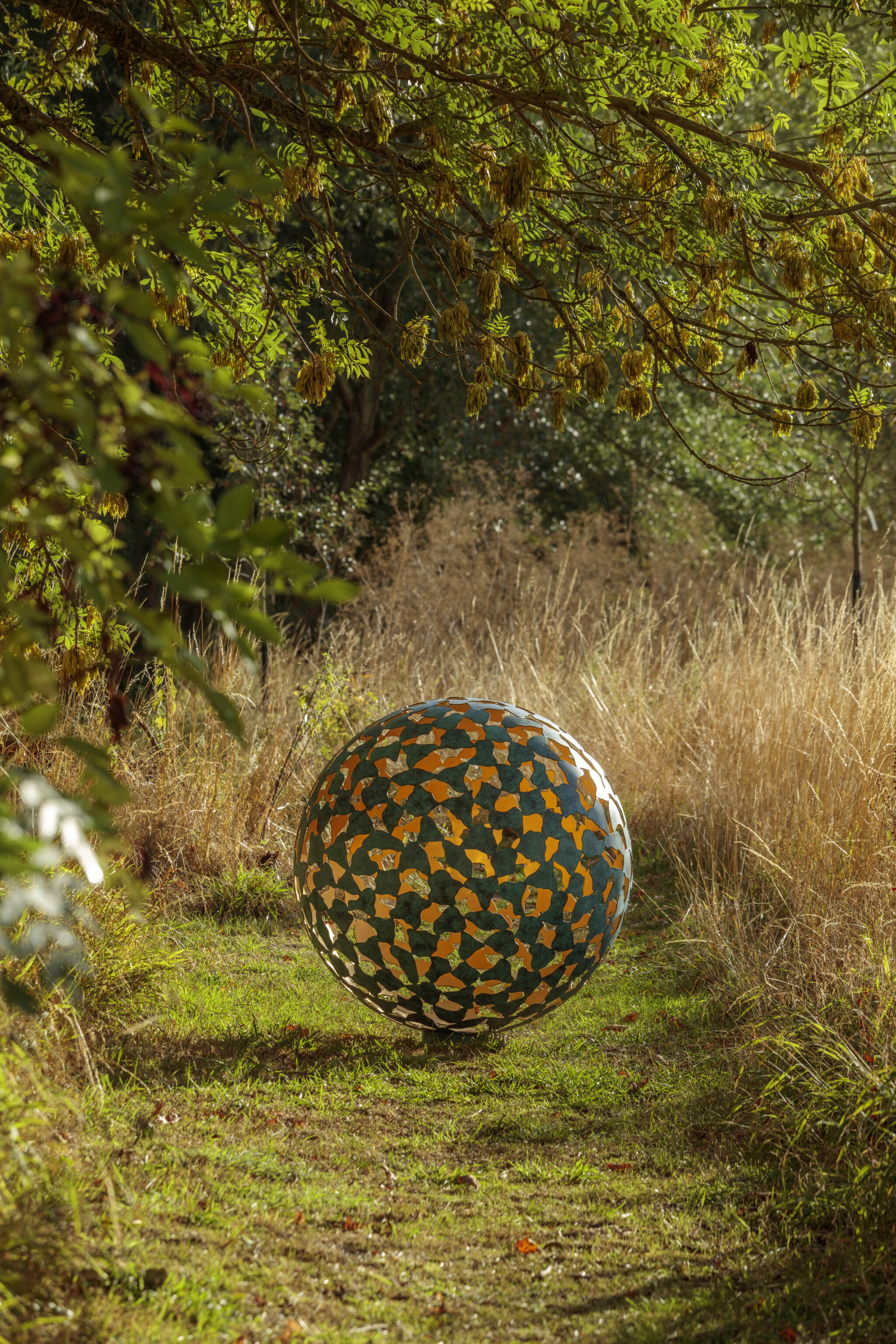 A Mantle II in bronze and gold leaf contemporary abstract sculpture sits on a grassy path amid tall grass and green trees.