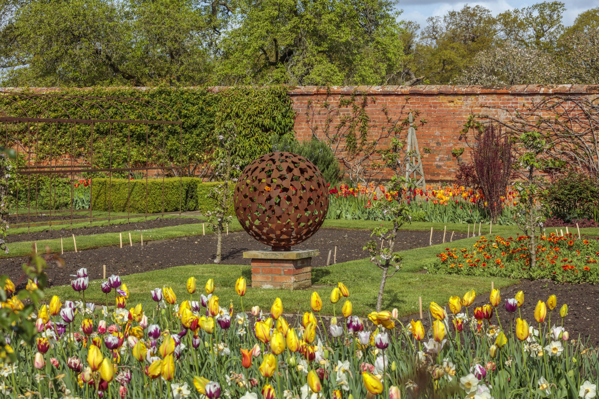 Garden with colourful tulips, a mantel oxidised steel sphere sculpture, and a brick wall in the background.