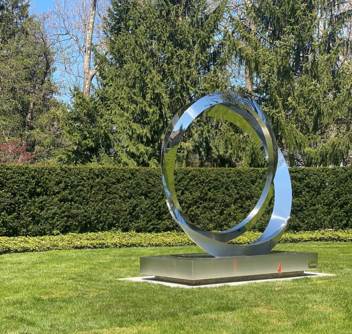 Shiny stainless steel eclipse sculpture on a grassy lawn with trees and hedges in the background.