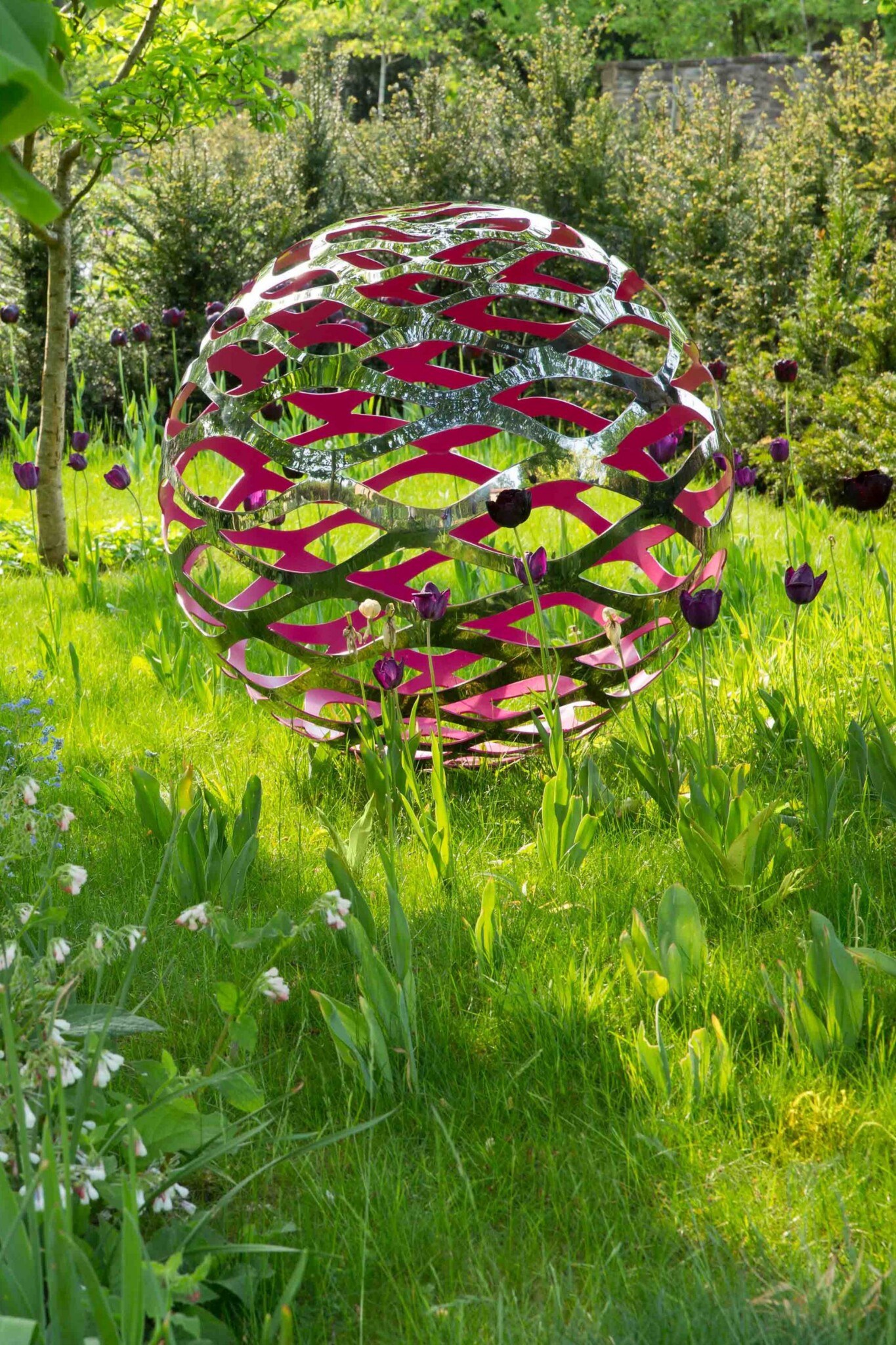 Silver metal sphere sculpture with a painted pink interior sits within a spring garden filled with blossom and tulips