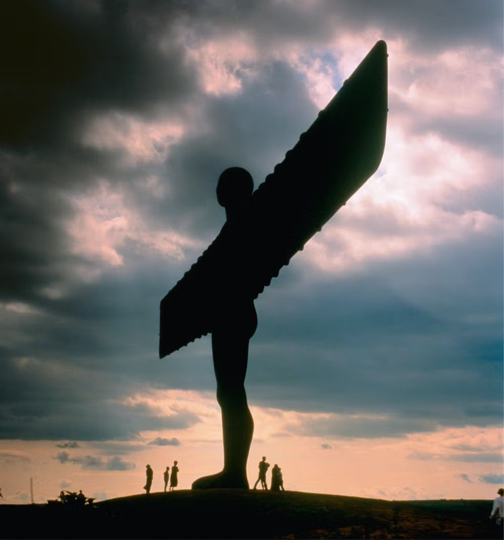 Silhouette of the Angel of the North outdoor art installation with people nearby, set against a dramatic sunset sky.