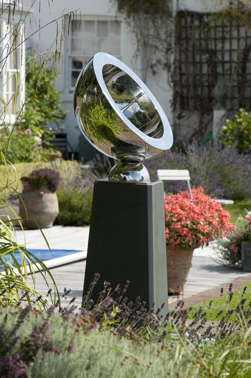 A shiny metal garden sculpture on a black plinth inspires courtyard garden ideas amid flowers and lush greenery.