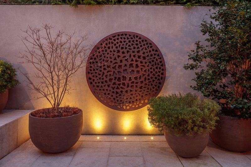 Large round metal art on a wall, lit from below for an inspiring courtyard garden idea with potted plants completing the look.