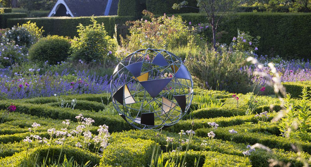 A contemporary abstract sculpture stands in a lush, flower-filled garden with manicured greenery.