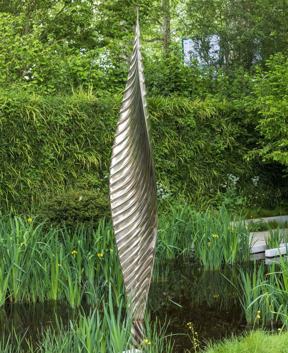 Tall, twisted contemporary abstract sculpture rises from a pond, surrounded by lush green plants and trees.