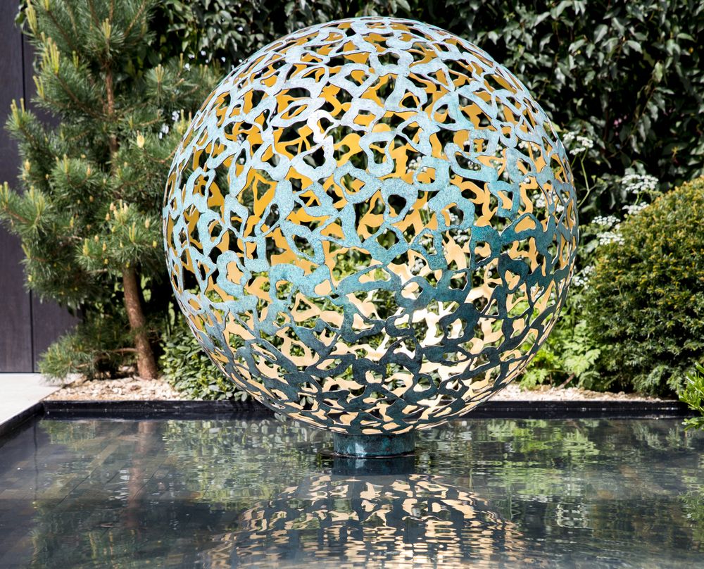 A contemporary abstract sculpture, this decorative metal sphere features a cut-out pattern on a reflective water garden surface.