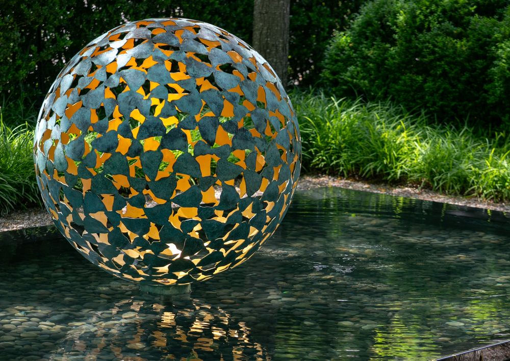 Contemporary abstract sculpture: spherical metal with cut-out shapes, reflected in a pond and surrounded by greenery.