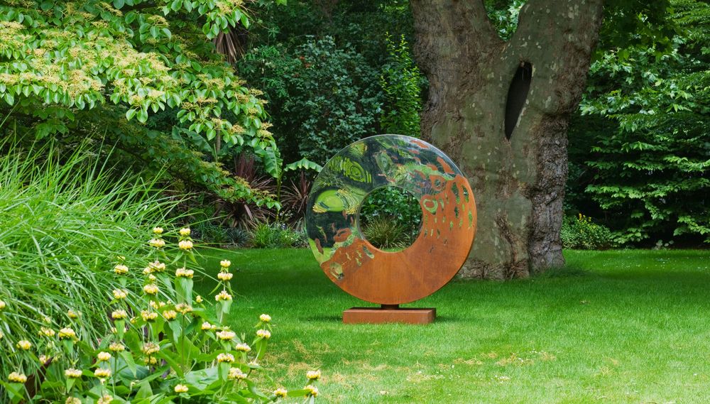 A contemporary abstract sculpture with a central hole stands on grass near a large tree in a lush garden.