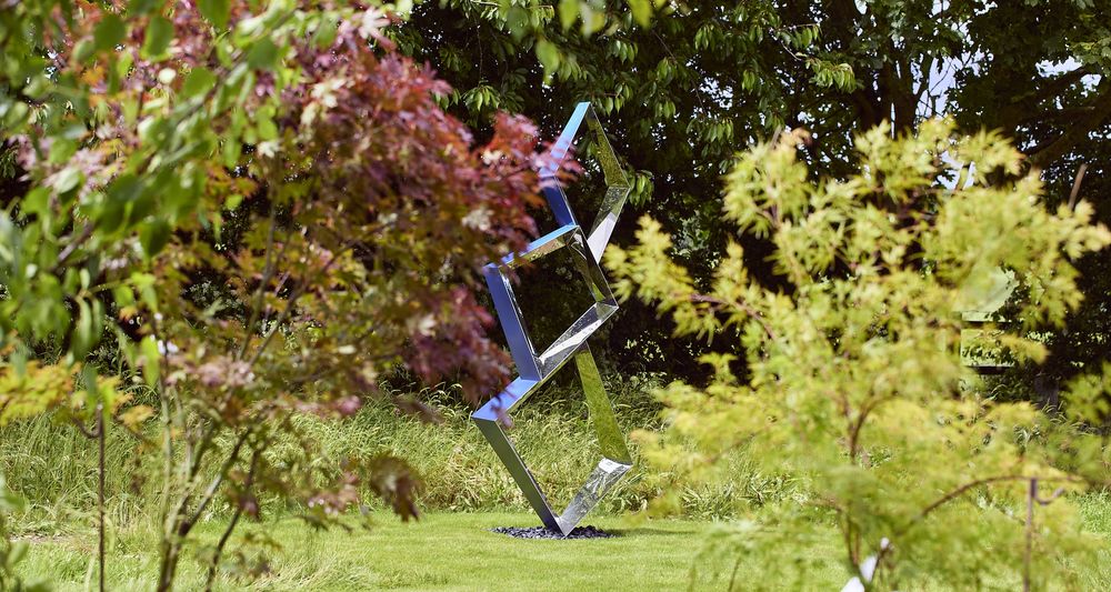 A shiny, contemporary abstract sculpture stands on a grassy lawn among green and red-leaved trees.