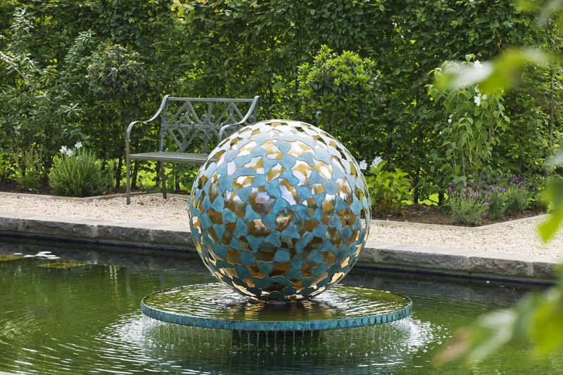 A large decorative metal sphere forms a custom water mantel atop a garden fountain, with a bench and green hedges behind.