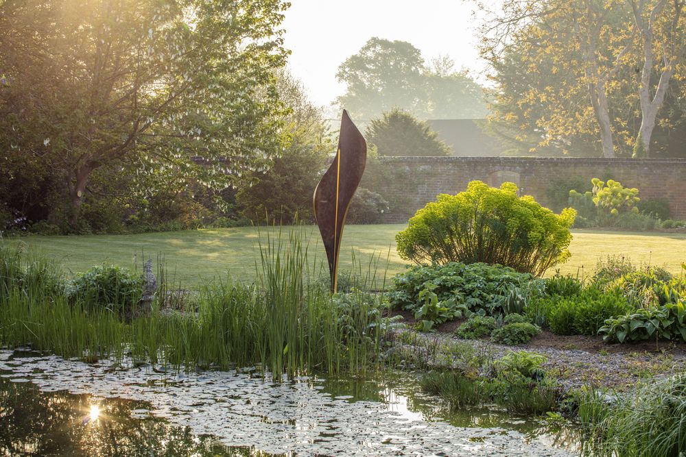 A serene garden scene at sunrise features a pond with waterlilies, lush plants, and an abstract metal sculpture that adds striking garden décor. Sunlight filters through trees, highlighting the manicured lawn and a brick wall in the background.