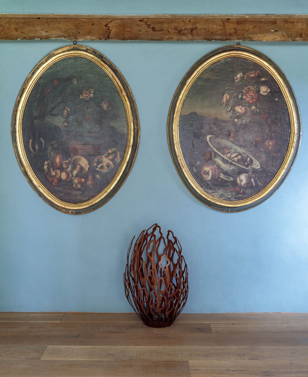 Two oval still life paintings hang above an alba floor sculpture in a stylish living room on a wooden table.