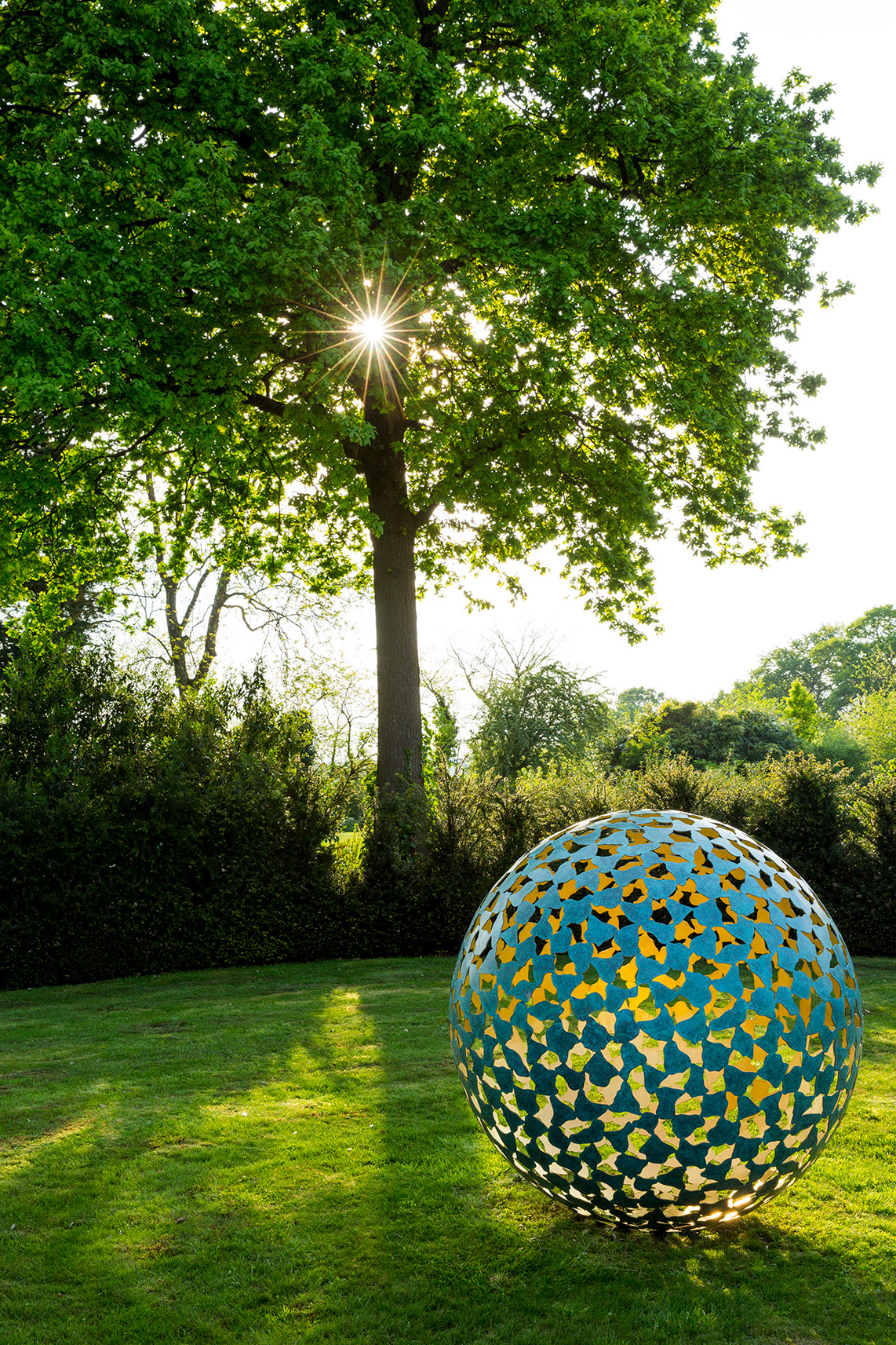 A large spherical sculpture, Mantle II in bronze and gold leaf, with butterfly-shaped cutouts stands on a grassy lawn. Sunlight filters through leafy trees, casting dappled light and shadows. The contemporary abstract scene feels serene and vibrant.