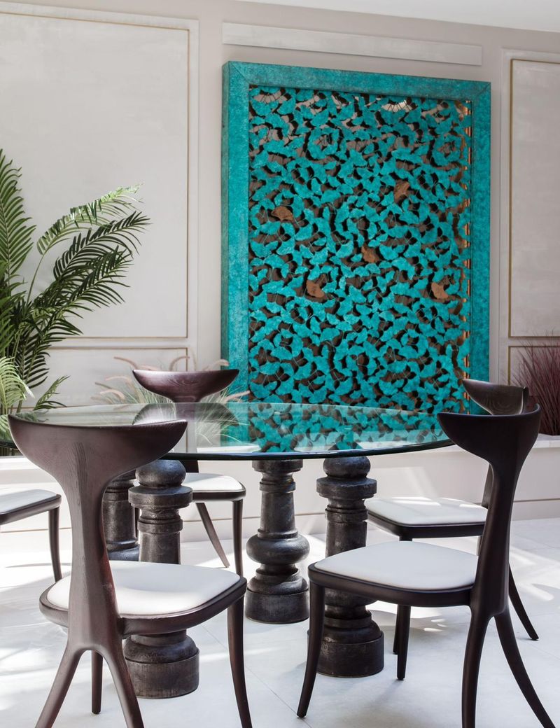Modern dining area with sculptural chairs, glass table, and stylish display sculptures at home against a turquoise wall panel.
