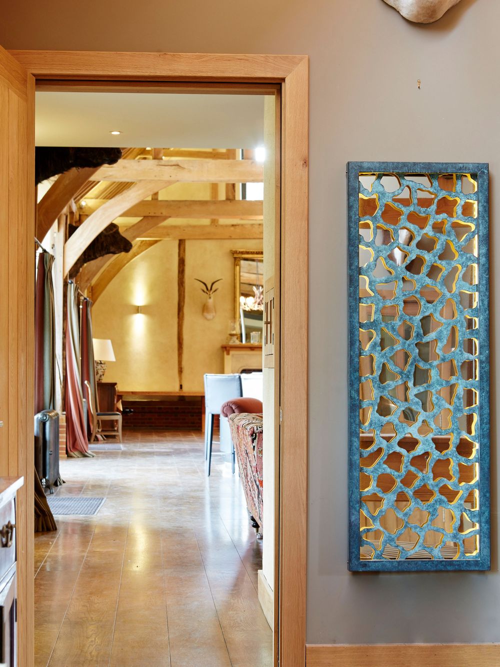 Modern hallway with wooden floors, a blue decorative wall mirror, and an indoor sculpture near a cozy living space with beams.