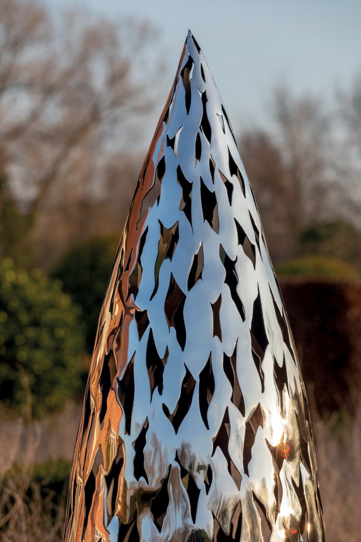 Shiny quill stainless steel sculpture with leaf-shaped cut-outs, set outdoors amid blurred trees.