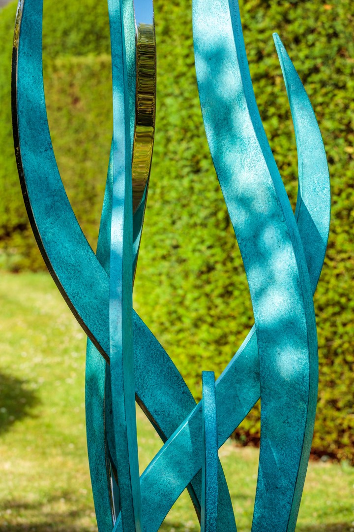 A modern hydra bronze and gold leaf contemporary abstract sculpture features turquoise blue, curved metal beams intertwining vertically, with a small golden reflective element nestled between two beams. The bright green hedge and lawn form a vibrant backdrop on a sunny day.