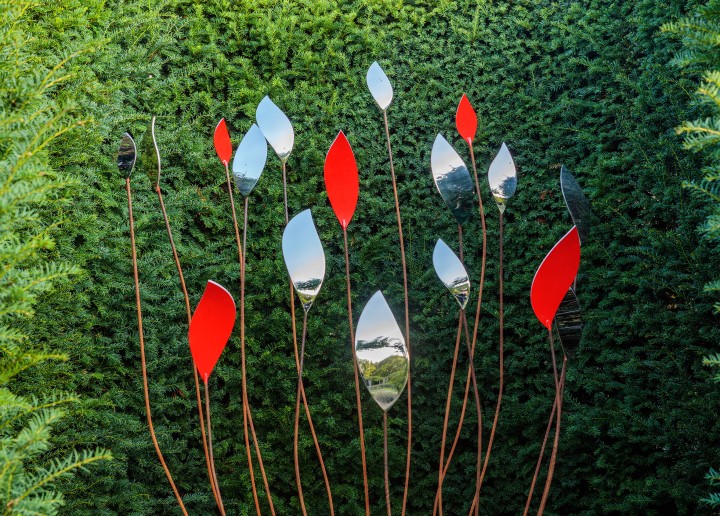 Tall stainless steel garden sculptures with red quiver shapes stand against a dense green hedge backdrop.