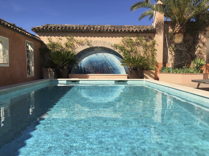 A serene outdoor pool with clear blue water is surrounded by beige walls, plants, and palm trees. At one end, a mosaic sundial accentuates the fountain’s blue arch, while sunlight reflects on the water under a clear sky.
