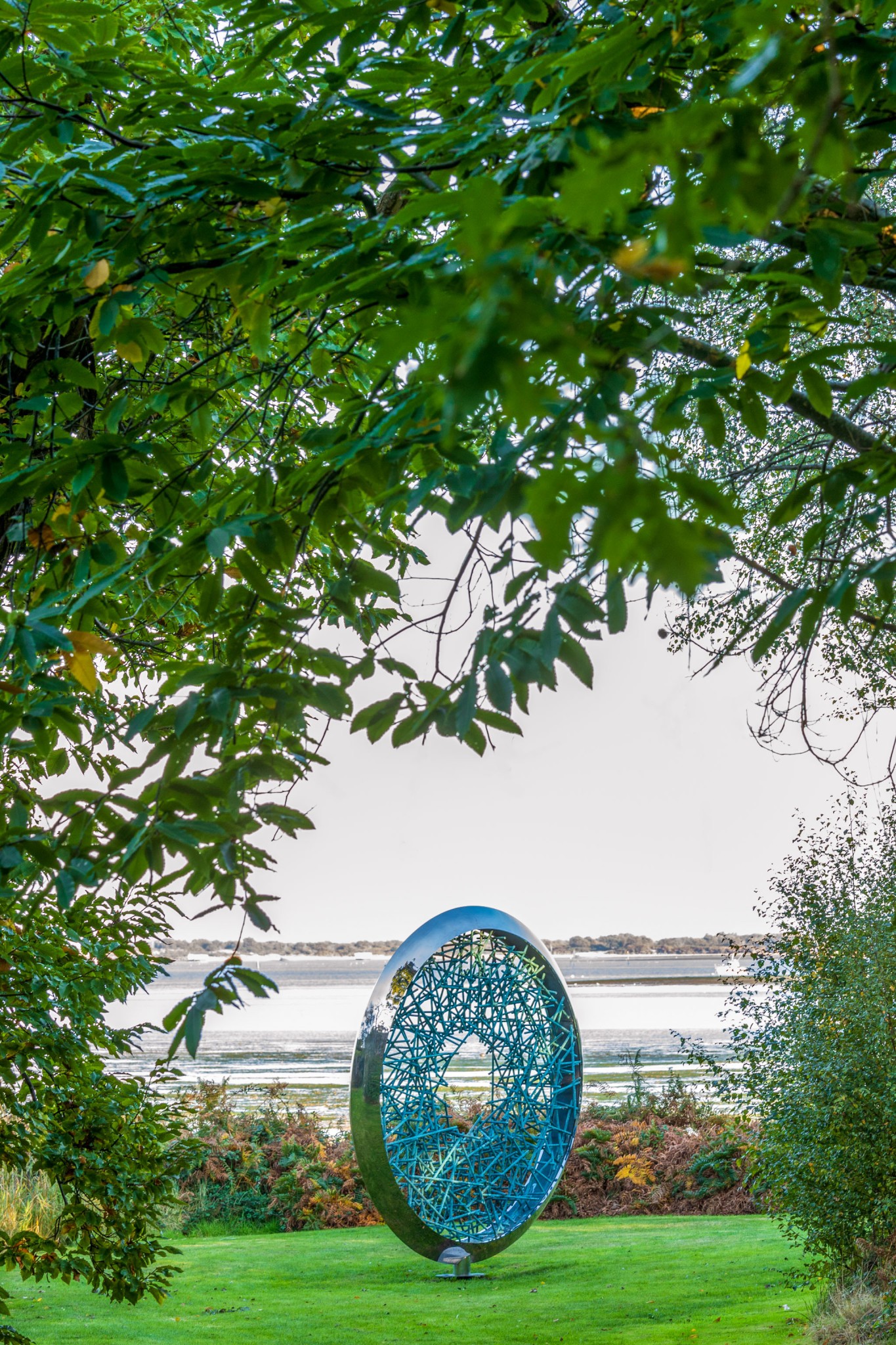 A large Ortus verdigris copper abstract sculpture stands on grass, framed by green trees, with water in the background.