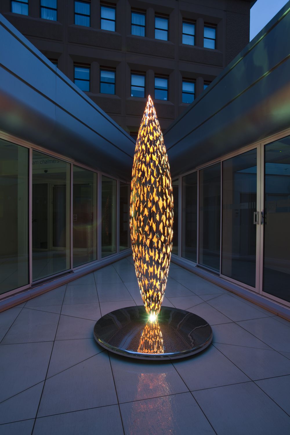 A tall, quill floor sculpture glows in a modern courtyard surrounded by glass walls at twilight.