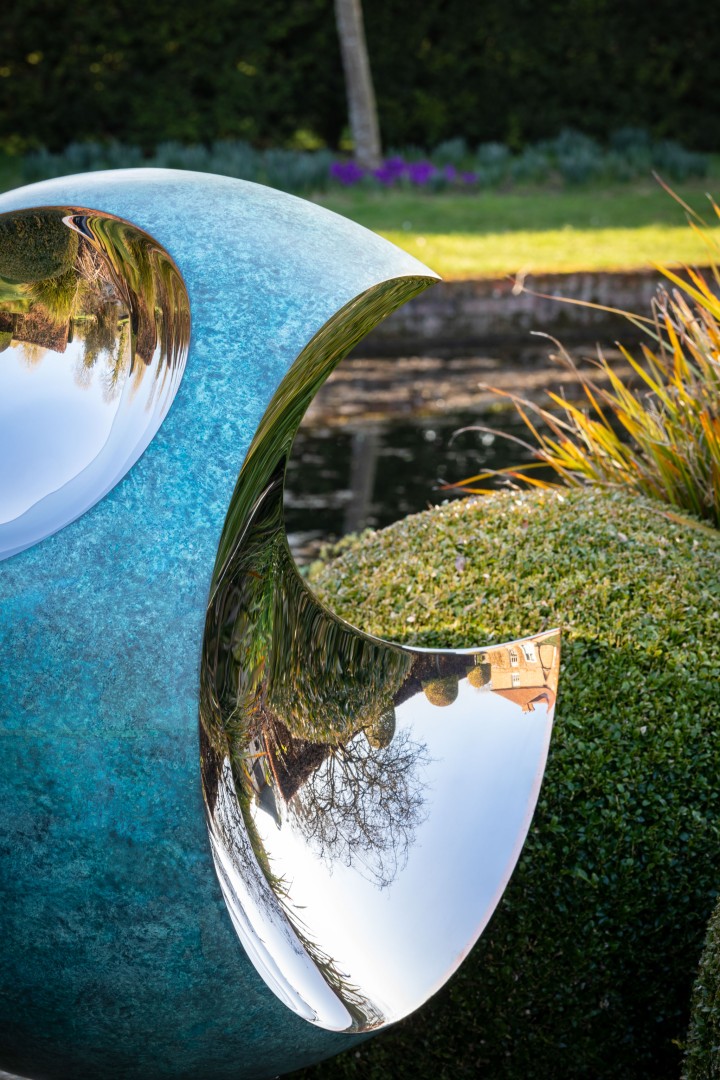A shiny Bite & Bite II stainless steel and copper abstract contemporary sculpture with a crescent-shaped cut-out stands outdoors near a pond, reflecting trees, bushes, and grass in its polished surface amidst manicured greenery and a blurred garden background.
