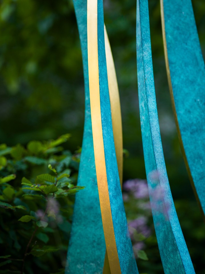 Tall, narrow teal and yellow ribbons evoke a hydra bronze and gold leaf contemporary abstract sculpture, rising vertically among blurred green foliage in soft natural light against a lush, leafy garden backdrop.