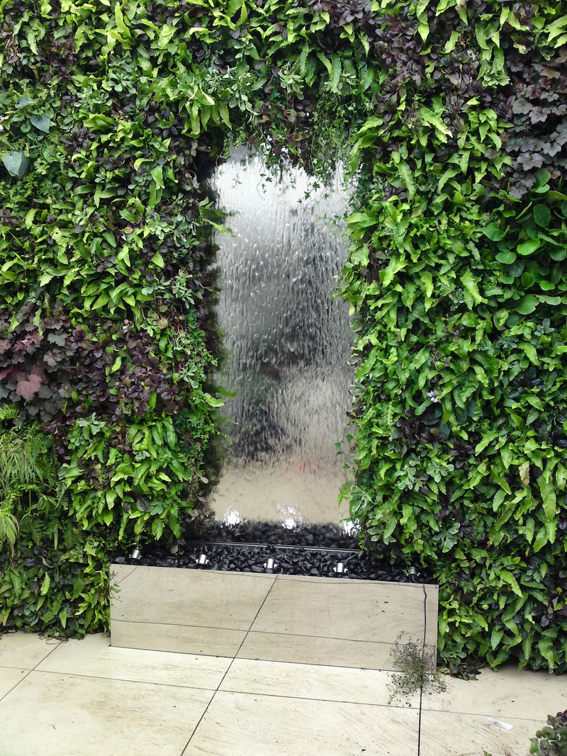 large stainless steel water feature with water cascading down a glass panel into a lush green living wall and stone basin