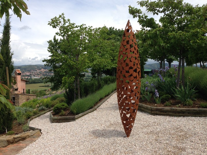 Quill-inspired oxidised steel contemporary sculpture stands on a gravel path in a lush garden with distant hills.