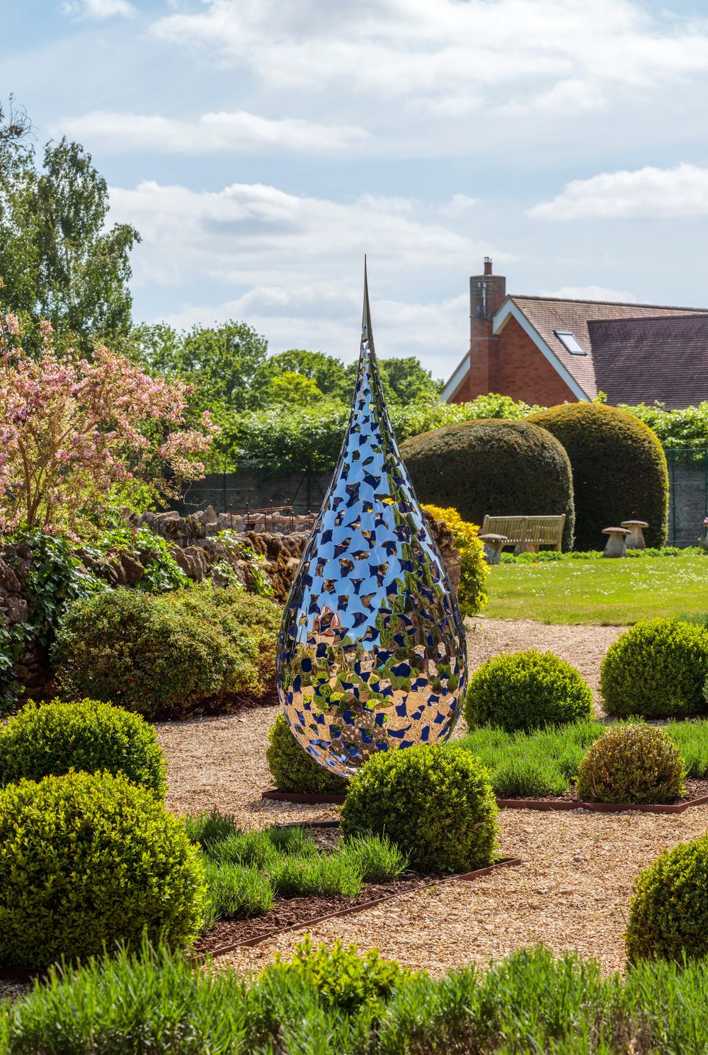 A shiny teardrop garden sculpture idea stands amid shrubs with a house in the background.