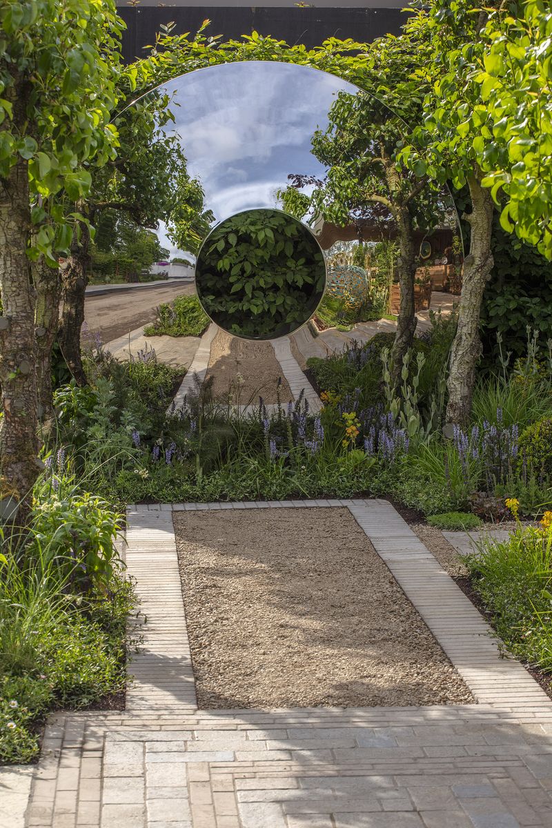 A circular mirror sculpture reflects trees and sky, inspiring courtyard garden ideas along a gravel path with flowers.
