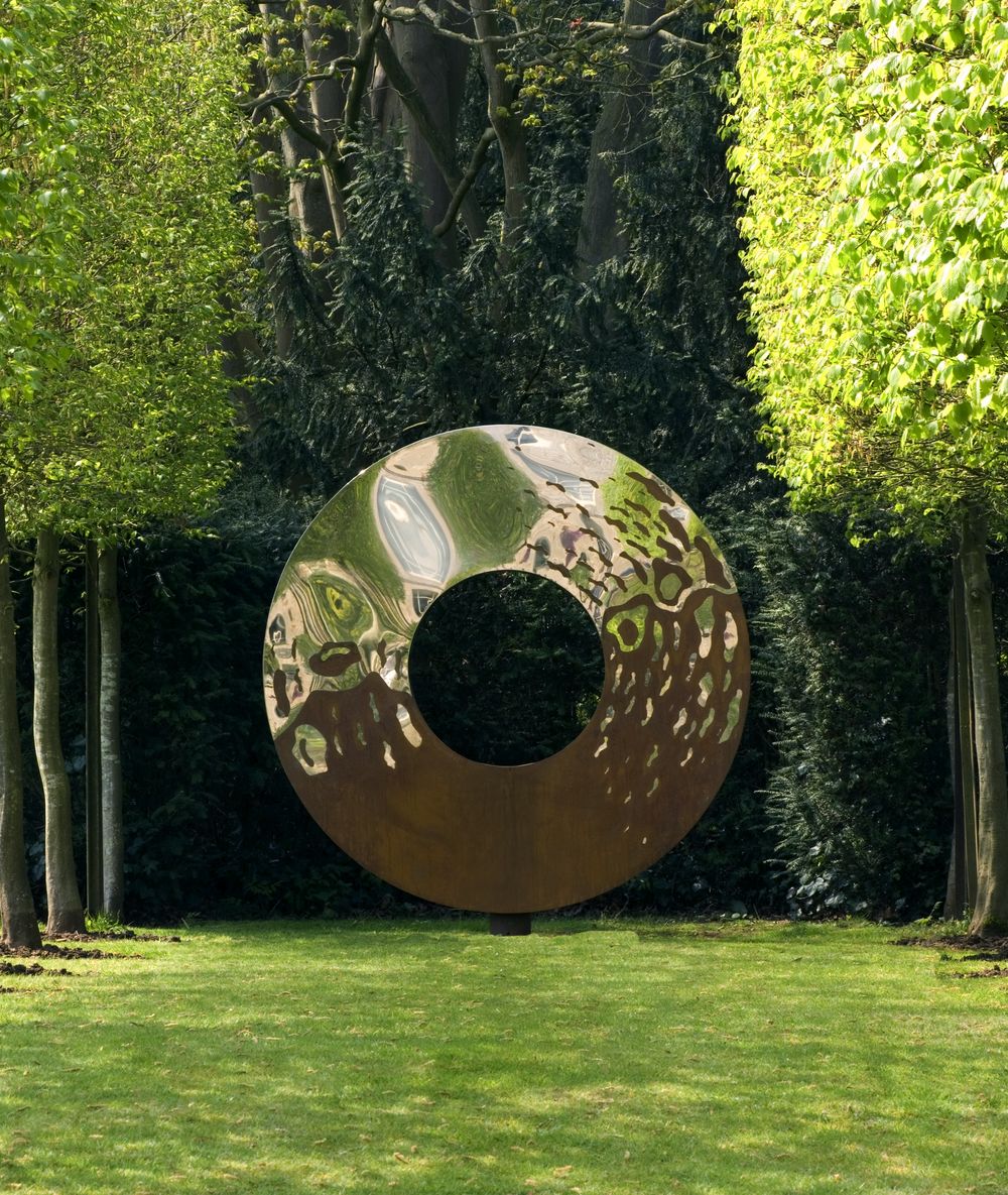 Large torus garden sculpture idea: circular metal art with a central hole, set on grass between lush green trees.