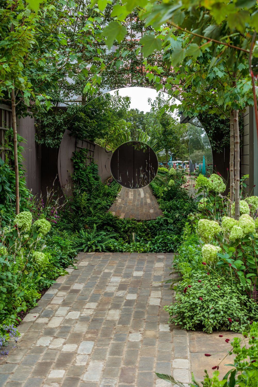 A paved brick path winds through lush green plants and white hydrangeas, leading to a striking Torus organic sculpture with a smaller circle at its base. Tall trees and wooden fences frame the walkway, creating a tranquil and inviting garden design.