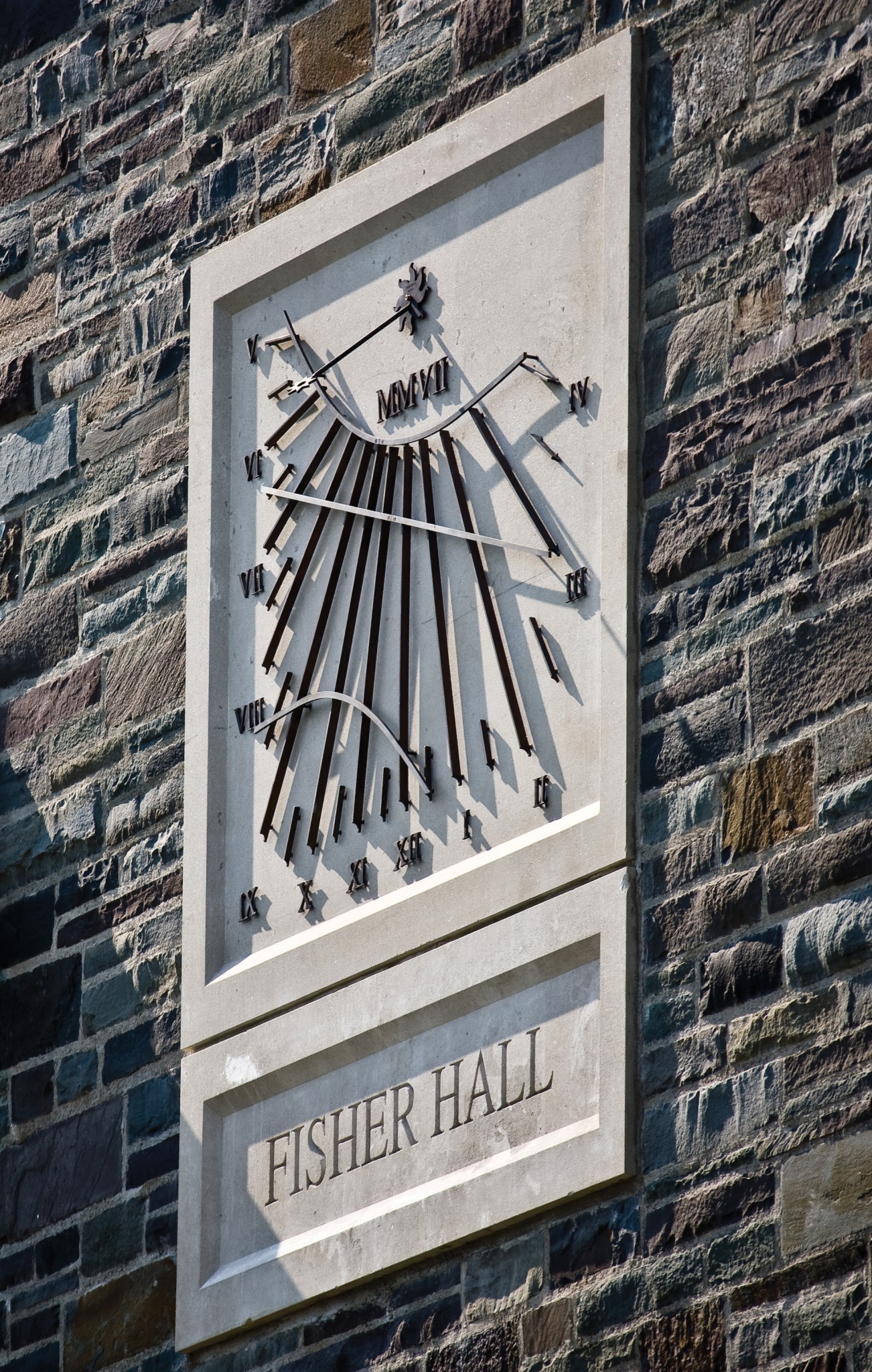 A stone wall with a rectangular Vertical Dial sundial featuring Roman numerals and metal rods casting shadows. Below the sundial, an engraved plaque reads FISHER HALL. The wall is made of multicoloured, irregularly shaped stones.
