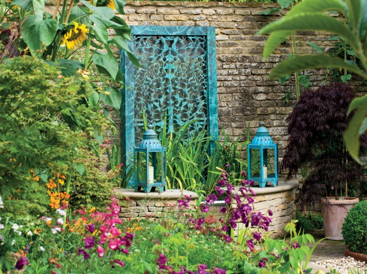 Lush garden with colorful flowers, stone wall, abstract Filigree Water Wall, and two blue lanterns with bronze filigree.