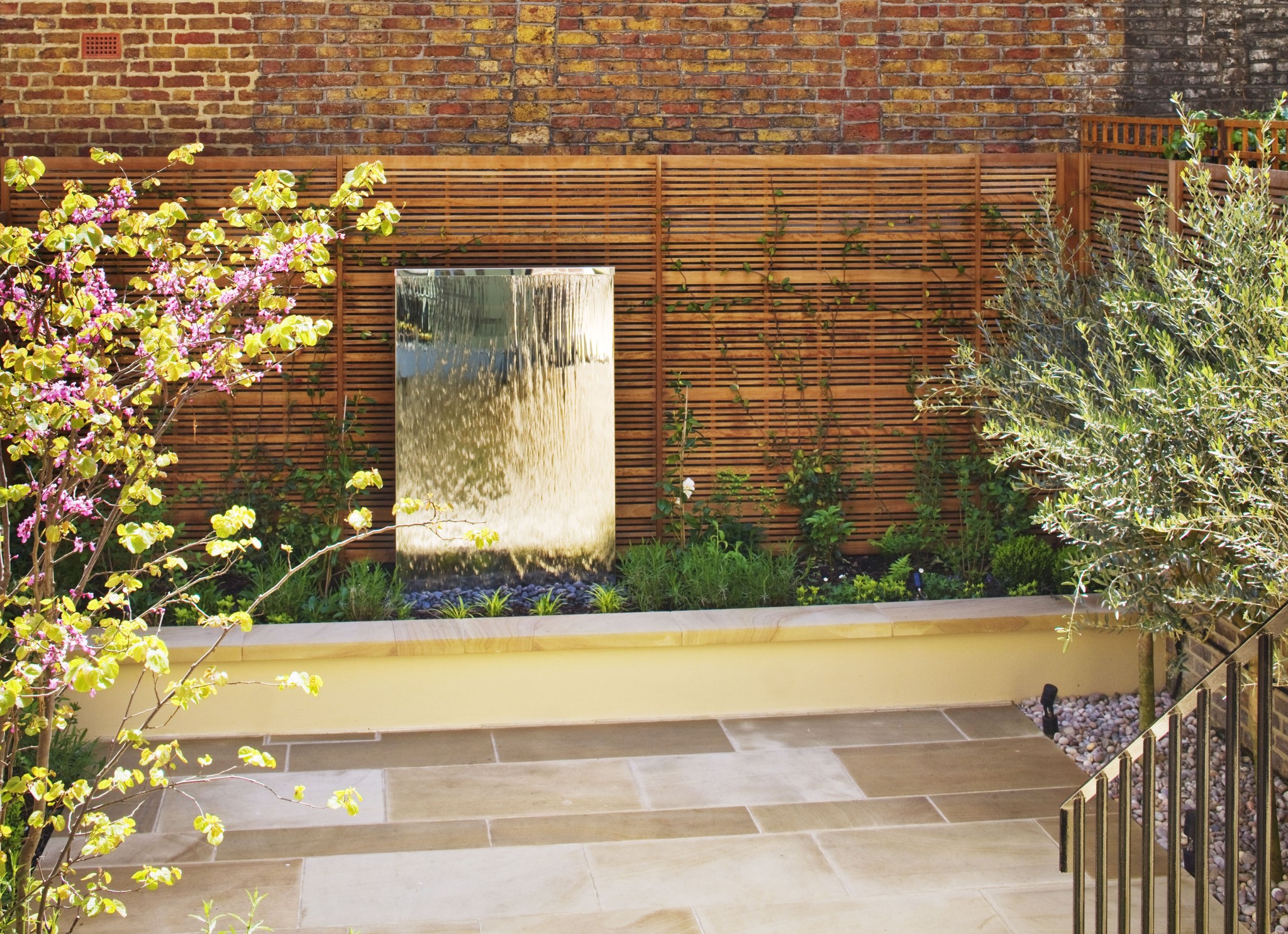 Small modern garden with stone patio, flowering plants, and a stainless steel water feature against a wooden fence.