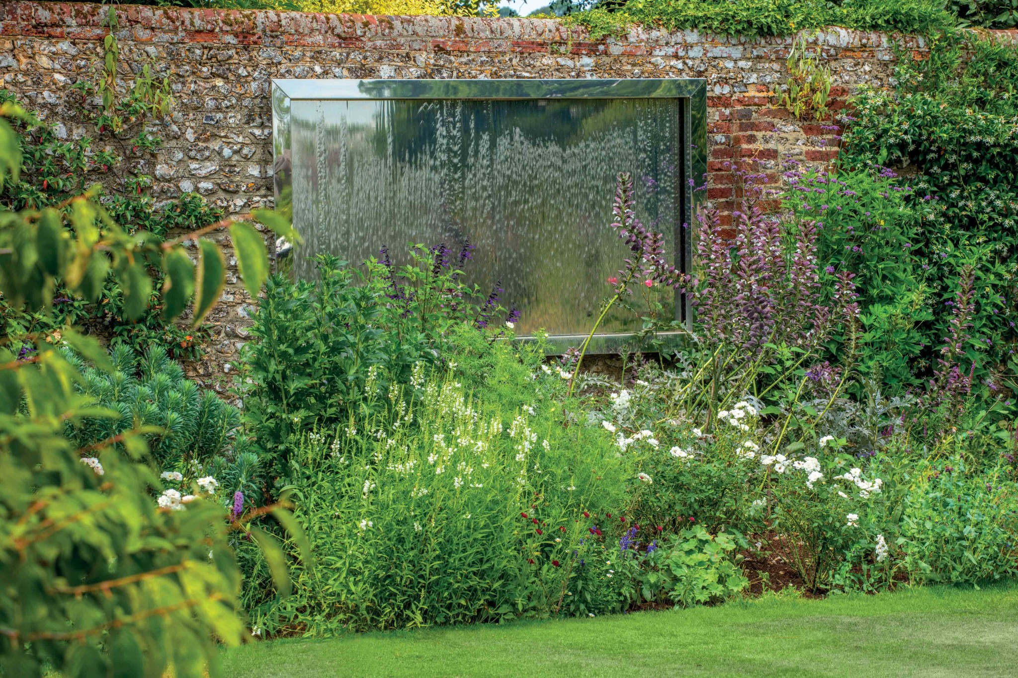 A lush garden with flowering plants in front of a brick wall and a striking stainless steel water feature.